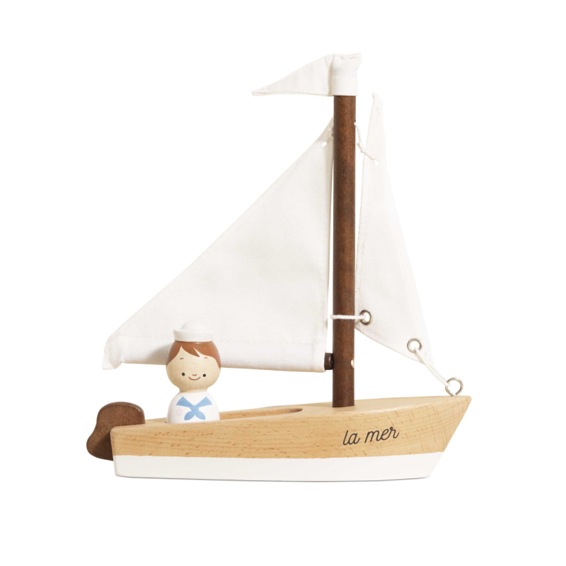 A wooden toy sailboat with white sails, a brown mast, and a small painted sailor figure on a white background. The hull is light natural wood with the text 'la mer' written on it.